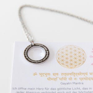 Könnte beinhalten: Silberfarbene Halskette mit rundem Anhänger und eingraviertem Text. Die Kette liegt auf einer weißen Karte mit goldenem geometrischen Design und Text in einer Fremdsprache.