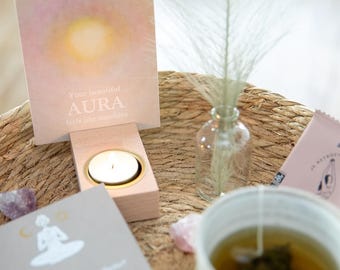 May include: A wooden candle holder with a lit tea light candle. A card with the text "Your beautiful aura feels like sunshine" is displayed on the holder. A wicker basket with a teacup, tea bag, and a small glass bottle with dried flowers is in the background.