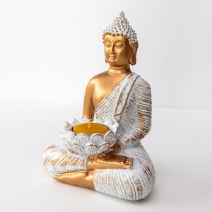 Könnte beinhalten: Eine sitzende Buddha-Statue in Gold und Weiß, die einen Lotusblüten-Kerzenhalter mit einer gelben Kerze hält. Die Statue weist detaillierte Schnitzereien auf. Der Kerzenhalter ist weiß mit goldenen Akzenten.