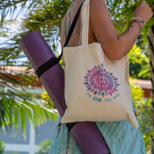 Könnte beinhalten: Eine Person trägt eine lila Yogamatte in einer beige Tragetasche mit dem Text "I'm OM my way." und einem farbenfrohen Mandala-Design. Die Tasche hat einen langen Riemen und wird über der Schulter getragen.