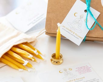 May include: A collection of yellow taper candles with white wicks, displayed with a gold candle holder and a small drawstring bag. The image includes a brown box and cards with the text "am mindful & focused". The candles are a warm, inviting color.