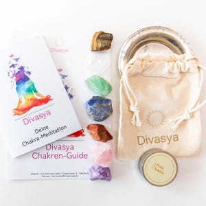 Könnte beinhalten: Ein Set aus sieben Chakra-Kristallen, darunter Amethyst, Rosenquarz, roter Jaspis, Lapislazuli, grüner Aventurin, klarer Quarz und Tigerauge. Die Kristalle sind in einer Reihe auf einer weißen Oberfläche angeordnet. Es gibt eine kleine Tasche, ein Booklet und einen kleinen Behälter mit dem Text "Palo Santo" darauf.