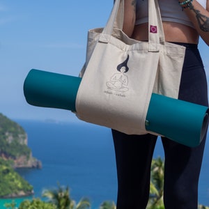 Puede incluir: Una persona lleva una bolsa de lona beige con una esterilla de yoga dentro. La bolsa tiene una ilustraci&oacute;n en blanco y negro de una persona meditando con el texto "inhale - exhale".