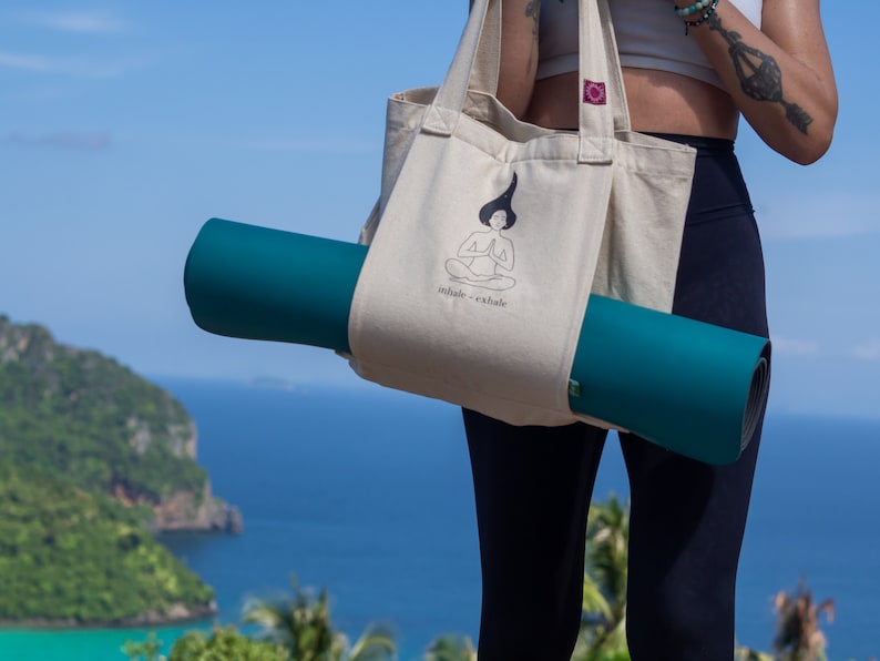 Puede incluir: Una persona lleva una bolsa de lona beige con una esterilla de yoga dentro. La bolsa tiene una ilustraci&oacute;n en blanco y negro de una persona meditando con el texto "inhale - exhale".
