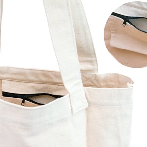 Puede incluir: Una bolsa de lona blanca con un bolsillo con cremallera en la parte delantera. La bolsa tiene dos asas largas para llevar.