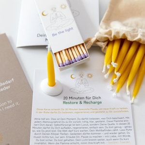 Könnte beinhalten: Eine Auswahl an Entspannungs- und Meditationsartikeln. Das Bild zeigt eine Streichholzschachtel mit dem Text "Be the light", eine gelbe Kerze in einem weißen Halter und mehrere gelbe Kerzen in einem beigen Beutel. Es gibt auch eine Karte mit Text.