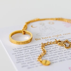 Könnte beinhalten: Goldkette mit einem Ringanhänger, auf dem "Be Here Right Now" steht. Die Kette liegt auf einem weißen Hintergrund mit Text in deutscher Sprache.