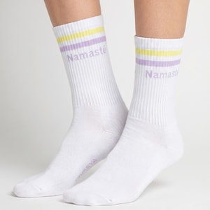 Könnte beinhalten: Weiße Sportsocken mit gelben und lila Streifen am oberen Rand. Das Wort "Namaste" ist lila auf der Seite jeder Socke aufgedruckt. Die Socken sind gerippt und aus weichem Material.