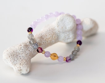 May include: A purple and grey beaded bracelet with a gold charm. The bracelet is on a white coral-like surface.
