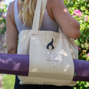 Puede incluir: Una bolsa de lona beige con una esterilla de yoga metida dentro. La bolsa tiene un gr&aacute;fico de una mujer meditando con el texto "inhale - exhale".
