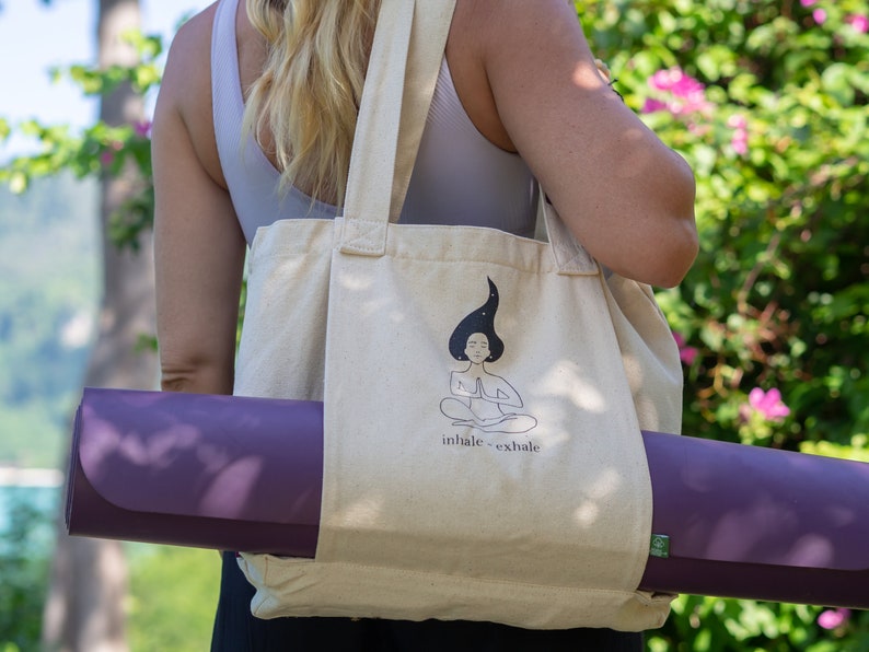 Puede incluir: Una bolsa de lona beige con una esterilla de yoga metida dentro. La bolsa tiene un gr&aacute;fico de una mujer meditando con el texto "inhale - exhale".