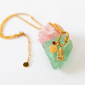 Puede incluir: Un collar dorado con múltiples colgantes, incluyendo un diseño de sol, una figura de deidad y una gema roja. El collar se exhibe sobre una piedra rosa y verde contra un fondo blanco.