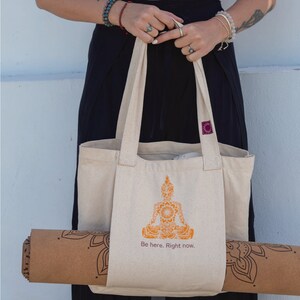 Puede incluir: Una bolsa de lona beige con una esterilla de yoga marr&oacute;n en su interior. La bolsa tiene un gr&aacute;fico de un Buda meditando con el texto "Be here. Right now." impreso en la parte delantera.
