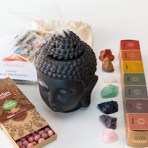 May include: A black Buddha head incense burner with a textured top, emitting smoke. The image includes a box of incense, a small bag, seven chakra stones, and boxes labeled "SEVEN CHAKRAS". The text "Divasya" and "Sagrada Madre" are visible.