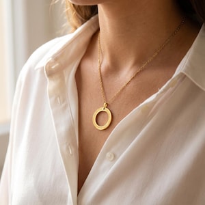Può includere: Una collana con pendente color oro con un design circolare. Il pendente pende da una delicata catena e poggia su una camicia bianca abbottonata. Il pendente circolare presenta un motivo inciso.