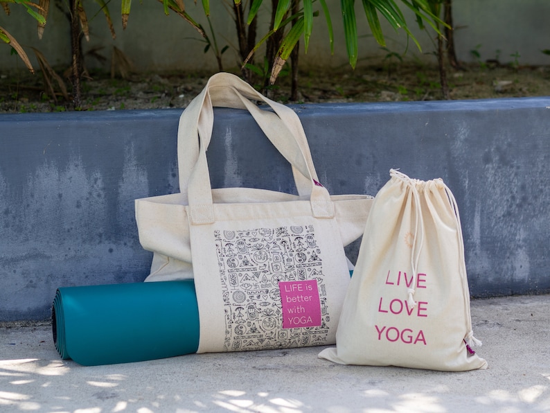 Puede incluir: Dos bolsas de lona blancas con texto rosa. Una bolsa tiene el texto "Life is better with yoga" y la otra bolsa tiene el texto "Live Love Yoga". Una esterilla de yoga azul verdoso est&aacute; delante de las bolsas.