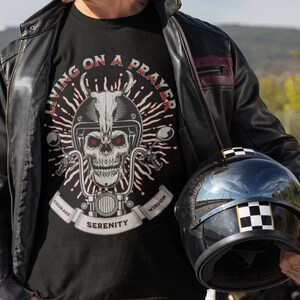 May include: Black t-shirt with a skull graphic wearing a motorcycle helmet. The graphic includes the text "LIVING ON A PRAYER" in red, with the words "COURAGE", "SERENITY", and "WISDOM" on a banner. A black motorcycle helmet with a checkered pattern is also visible.