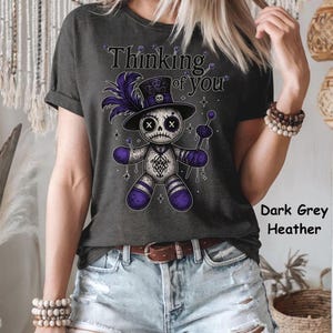 May include: Dark grey heather t-shirt featuring a voodoo doll graphic with a top hat and the text "Thinking of You". The doll is white with purple accents and holding a purple wand. The shirt is worn with denim shorts.