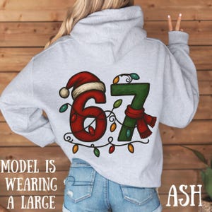 May include: Ash gray hoodie with a festive Christmas design. The number "67" is decorated with a Santa hat, scarf, and string lights. The text "MODEL IS WEARING A LARGE" is printed on the front.