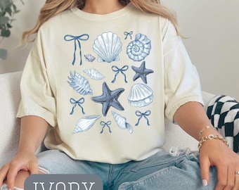 Op de afbeelding: Ivoor kleurig T-shirt met blauwe illustraties van schelpen en strikken. Het ontwerp bevat verschillende schelpen en zeesterren, met het woord "IVORY" onderaan gedrukt. Het shirt heeft een relaxte pasvorm.