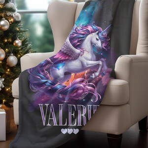 May include: A black blanket draped over a cream-colored armchair. The blanket features a majestic white unicorn with a flowing purple and pink mane and tail, and wings. The name "VALERIE" is printed on the blanket, along with three white hearts.