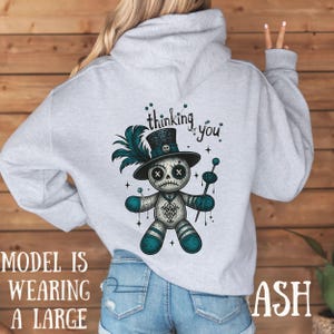 May include: Grey hoodie with a voodoo doll graphic. The design features a doll with a skull face, a top hat, and teal details. The words "thinking of you" are above the doll. The model is wearing a large.