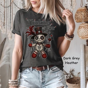 May include: Dark grey heather t-shirt featuring a voodoo doll graphic with the text "Thinking of You." The doll has a skull face, a top hat, and red accents. The shirt is a crew neck style.