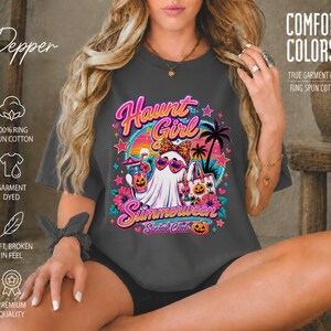 Puede incluir: Una camiseta gris oscuro de manga corta con un estampado gr&aacute;fico de un fantasma con gafas de sol en forma de coraz&oacute;n y un lazo de leopardo. El dise&ntilde;o incluye palmeras, una puesta de sol, flores tropicales y el texto Haunt Girl Summerween Social Club.