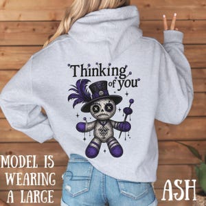 May include: Light gray hoodie with a graphic of a voodoo doll wearing a top hat and holding a staff. The text "Thinking of you" is above the doll. The doll is purple, black, and white. The word "ASH" is at the bottom right.