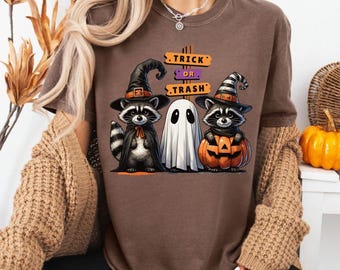 Op de afbeelding: Bruin t-shirt met een Halloween-ontwerp. Het toont twee wasberen met heksenhoeden en een spook, met één wasbeer in een cape en de andere met een uitgesneden pompoen. De tekst op het shirt luidt "Trick or Trash".
