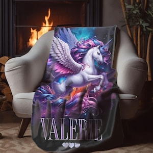 May include: A plush throw blanket featuring a majestic unicorn with flowing mane and wings, in shades of purple, pink, and blue. The blanket has the name "VALERIE" printed on it, with three hearts below. The blanket is draped over a white armchair.