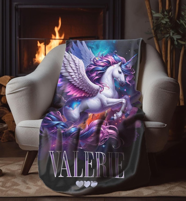 May include: A plush throw blanket featuring a majestic unicorn with flowing mane and wings, in shades of purple, pink, and blue. The blanket has the name "VALERIE" printed on it, with three hearts below. The blanket is draped over a white armchair.