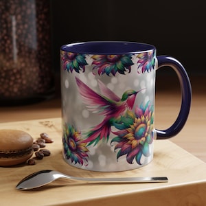 May include: A white ceramic mug with a blue rim and handle. The mug features a colorful hummingbird flying over a field of sunflowers. The background is a silver shimmer.