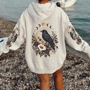 Könnte beinhalten: Ein hellgrauer Kapuzenpullover mit einem großen Grafikdruck auf dem Rücken und den Ärmeln. Das Motiv zeigt einen schwarzen Raben auf einer goldenen Mondsichel, umgeben von violetten Blumen, Beeren und Mondphasensymbolen.