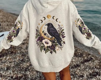 Peut inclure: Un sweat à capuche gris clair avec un grand motif imprimé au dos et sur les manches. Le dessin central représente un corbeau noir posé sur un croissant de lune doré, entouré de fleurs violettes, de baies et de symboles lunaires.