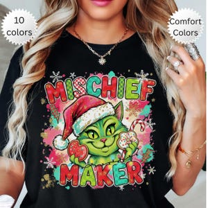 May include: Black t-shirt featuring a green cat in a Santa hat, holding a candy cane and biscuits. The words "MISCHIEF MAKER" are in festive colours, with snowflakes and Christmas decorations. A seasonal design.