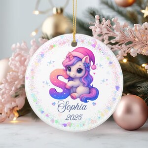 Puede incluir: Adorno navideño redondo con un unicornio de dibujos animados que lleva un gorro de Papá Noel rosa y sostiene la letra "S". El adorno tiene el nombre "Sophia" y el año "2025". Es blanco con una cuerda dorada para colgar.