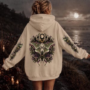Op de afbeelding: Een beige hoodie met een grote afbeelding op de achterkant van een groene nachtvlinder omringd door maanfasen, paarse paddenstoelen en bladeren. De mouwen zijn voorzien van bijpassende botanische illustraties en nachtvlinders.