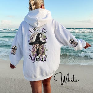 May include: A white hooded sweatshirt featuring a graphic on the back and sleeves. The back design shows a witch in a black hat with long hair, a full moon, bats, and the text A Wee Bit Wicked. Smaller versions of the witch graphic appear on each sleeve.