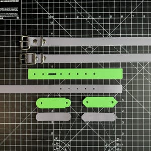 May include: A set of two dog collars, one gray and one green, with matching green and gray buckle straps. The collars are laid out on a black cutting mat with grid lines.