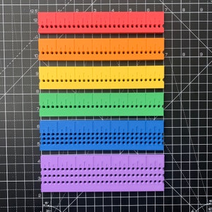 May include: Six colourful plastic rulers with holes along the edges. The rulers are red, orange, yellow, green, blue, and purple. Each ruler has markings for inches and fractions of an inch.