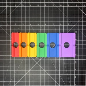 May include: Six colourful plastic blocks with a round hole in the centre. The blocks are arranged in a row from left to right: red, orange, yellow, green, blue, and purple.