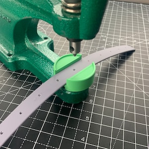 May include: A green leather craft punch with a metal base and a green plastic die is being used to punch a hole in a grey leather strap. The strap is lying on a black and white grid.