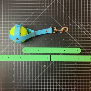 May include: A blue leather dog leash with a tennis ball attached to the end. The leash has a metal clip and two green straps with the letters "D" and "H" on them.