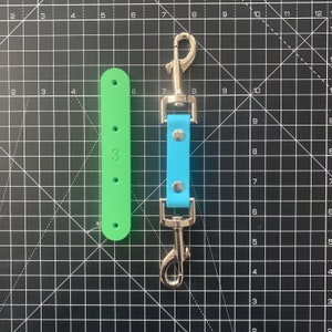 May include: A light blue leather dog lead connector with silver-tone metal fixings. A green, rectangular piece with the number "3" is next to the lead connector. The background is a black cutting mat with white grid lines.