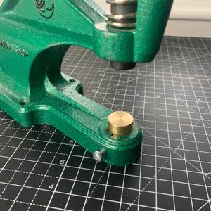 May include: A green metal hand press with a brass die. The press is sitting on a black grid surface.