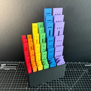 May include: A rainbow-colored set of plastic building blocks in a black storage box. The blocks are arranged in a graduated size order, with the largest blocks at the back and the smallest blocks at the front. The blocks are all different colors, including red, orange, yellow, green, blue, and purple.