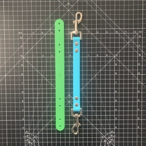 May include: Two leather dog leads, one green and one blue, with silver clasps. The leads are on a black grid background.