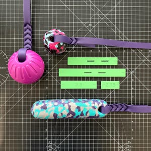 May include: A collection of dog toys and accessories, including a purple and pink ball, a camouflage-patterned chew toy, and green measuring tools. The toys are connected to purple straps. The green tools have measurements of 3.5 and 2.75 inches.