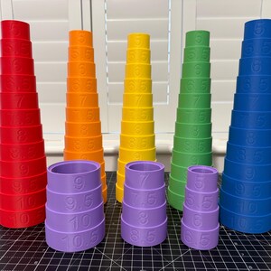 May include: A set of multi-coloured graduated cylinders in red, orange, yellow, green, and blue. Each cylinder is marked with numbers from 5 to 10.5, representing measurements. The cylinders are displayed on a black surface.